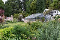 View over herbaceous perennial border to a nursery glasshouse and a small outbuilding, mature trees beyond