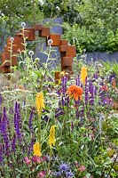 Yellow Kniphofia 'Tawny King' red-hot poker, purple Salvia sylvestris 'Mainacht', Echinops  and Eryngium planum sea holly with Echinacea  in front of 'Honesty' sculpture in Corten steel by Jill Clarke  - The Lower Barn Farm Outdoor Living Garden - RHS Hampton Court  Palace Garden Festival 2019