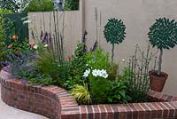 Curving, brick edged, raised bed planted with cosmos, roses, salvias, catmint and Verbena bonariensis. Behind are murals of potted bay trees on wall.