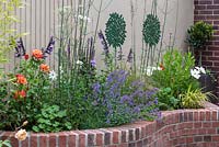 Dull wall enhanced with murals of potted bay trees. Below, curving, brick edged raised bed planted with cosmos, ammi, roses, salvias, catmint and Verbena bonariensis.