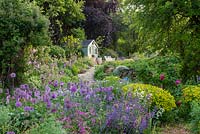 View through Allium 'Purple Sensation', Persicaria bistorta 'Superba', euphorbia, ragged robin, orlaya and catmint, to a paved path that ascends to a summerhouse beneath a copper beech.
