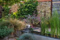 A salvaged galvanised water trough is converted into a water feature, and planted with irises and water lilies.  Beyond, a raised bed of euphorbia, cardoon and Erysimum 'Bowles Mauve'. Climbing hydrangea 