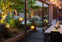 As night falls, garden lighting illuminates the covered dining area and planting beneath the acer, whilst the fire pit is reflected in the house windows.
