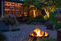 As the sun goes down, a flaming fire pit in the foreground adds to the garden lighting, with uplighters beneath an Acer palmatum and a string of bulbs above the dining table