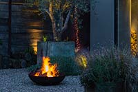 A blazing fire pit on gravel in a contemporary courtyard garden. Behind, a string of lights hang from an olive tree planted in an old galvanised water tank.