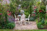 A small gravel courtyard with seating area surrounded by roses, poppies, astrantias and  Erysimum 'Bowles Mauve'.