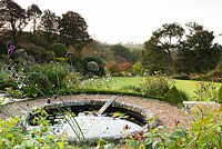 Circular pond edged with stone containing Nymphaea - Waterlily - and board for hedgehogs to climb out. Gravel path around pond offers views of garden lawn edged with borders and countryside views beyond
