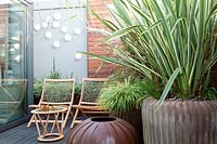 Wooden chairs are surrounded by potted planted, including Phormium tenax 'Variegatum' and Carex buchananii in modern terrace garden. 