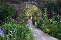 Gravel pathway through gate set in arch of old brick boundary wall by flowering summer borders. 