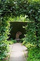 Path through arch in hedge framing sculptural stone seat by Taxus hedge 