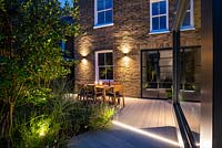 Wooden path leading to the illuminated dining area and house surrounded by borders with Gaura lindheimeriÂ 'Sparkle White', Salvia nemorosaÂ 'Caradonna', Miscanthus sinensisÂ 'Morning Light' and Osmanthus burkwoodii