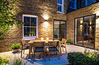 Night view across illuminated decked dining area towards brick house, with wooden dining table and chairs 