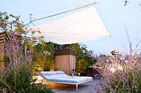 Large sun bed stands on terrace surrounded by Agapanthus, Perovskia atriplicifolia and Rosmarinum prostratrum, enhanced by garden lighting at dusk. 