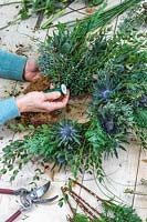 Woman attaching bundle of foliage and flowers to winter wreath with floristry wire. 