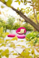 Relaxing area on a terrace viewed through shrubs. Mixed planting screens bright pink chair and cushions