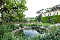 Circular pond with Rosa - Rose - bed at the centre of a formal garden, beyond rows of Citrus - Lemon - trees in pots and house 