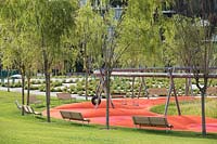 View of public park with young trees, benches and playground surrounded by planting and grass
