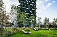 Public park with seating on grass with perimeter of trees and meadow. Behind  the Bosco Verticale - Vertical Forest - by architect Stefano Boeri 