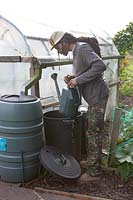 A man collecting water from a plastic dustbin linked to a water butt collecting rainwater off pipe on homemade polytunnel 