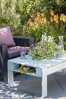 Wooden pallet table with integral sunken planter with summer planting, on slate patio in summer, accompanied by outdoor chairs.