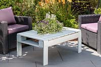 Wooden pallet table with integral sunken planter with summer planting on slate patio in summer accompanied by outdoor chairs.