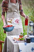 Woman wearing floral apron adding fresh fruit and drink to picnic table