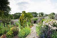 Curving, textured stone path leading between mixed borders including roses, day lilies, salvias and poppies toward a seating area 