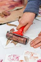 Woman rolling leaves painted with metallic spray paints onto paper bags using a lino roller. 