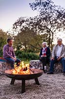 Lit firepit at dusk with people sitting on gabion benches 