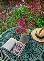 Still life with vase of cut Centranthus ruber - Red Valerian flowers, sun hat and insect book on garden table. 