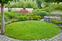 Small circular lawn with circular pond and perennial border beyond, edged with setts