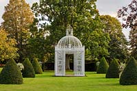 A white ornate metal gazebo flanked by clipped yew cones, Taxus baccata, and stone urns on pedestals with displays of silver-leafed Astelia chathamica. Behind it is the towering 'Queen Elizabeth Oak' - Quercus robus.