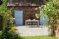 Graveled courtyard area with dining furniture and backdrop of outbuildings, in foreground Rosa - Climbing Rose - against a brick wall