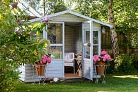 Wooden painted summerhouse with open door and view to comfortable furniture, pots with pink Hydrangea either side of door