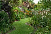 Views through a half-acre country garden, along a wide curving grass path, to borders of summer herbaceous perennials and pots of dahlias.