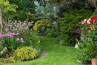 Views through a half-acre country garden past borders of summer herbaceous perennials and pots of dahlias, along a wide curving grass path to a bench in the shade of a tree.