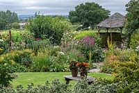 A wooden gazebo is flanked by borders of summer herbaceous perennials including mallows, lythrum, linaria, verbascum and phlox, with glimpses of Blackmore Vale beyond. 