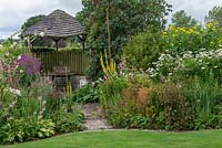 A wooden gazebo is flanked by borders of summer herbaceous perennials including mallows, lythrum, linaria, verbascum, rudbeckias, shasta daisies, liatris and phlox.