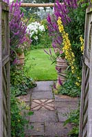 A rustic gateway frames a view across a path and past clumps of purple loosestrife to a lawn and clump of white phlox in far border.
