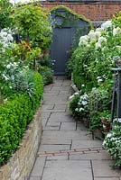 View along paved path to garden shed, passing raised beds edged in box hedging, planted with white agapanthus, cosmos, hydrangeas, geraniums and roses.