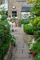 View up paved path to garden house, passing raised beds edged in box hedging, planted with white agapanthus, cosmos, hydrangeas, geraniums and roses. Tall table with steel dish of succulents.