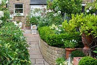 View up paved path to garden house, passing curving, brick raised bed edged in box hedging, planted with white agapanthus, fuchsias, cosmos, hydrangeas and roses.