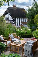 View over seating area to yew-edged beds of tall perennials, ornamental grasses and Cosmos, thatched cottage beyond