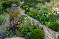 A bird's eye view of a contemporary garden design, a diagonal brick path separates a gravel garden from beds of perennials and grasses, interspersed with Taxus - Yew ball topiary 