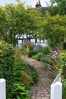 A brick path from gate to cottage, beds either side with multi-stemmed Amelanchier lamarchii underplanted with perennials