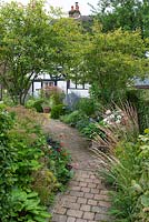 A brick path cuts diagonally through the garden, passing by two multi-stemmed Amelanchier lamarckii, to cottage