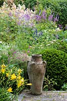An empty urn beside a Taxus - Yew - topiary dome, beyond a backdrop of hardy Geranium, Agastache, Veronicastrum virginicum and Hemerocallis - Daylily