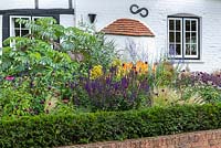 View over a boundary wall and hedge to a full border with: Melianthus major, Salvia, Kniphofia, Stipa and Allium, old cottage behind