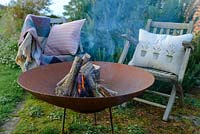 A lit fire bowl made from rusted corten steel,  wooden garden chairs with a wool rug and  ushions by a large rosemary bush.