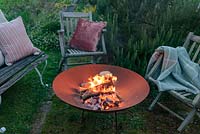 A lit fire bowl made from rusted corten steel, wooden garden chairs and a bench with wool rugs and  cushions by a large rosemary bush.